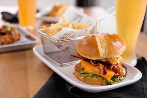 A close-up of a burger with bacon, cheese, and lettuce, served with fries and a drink in the background.
