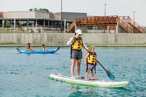 A woman and a child paddleboarding on a calm lake, with others in the background enjoying water activities.
