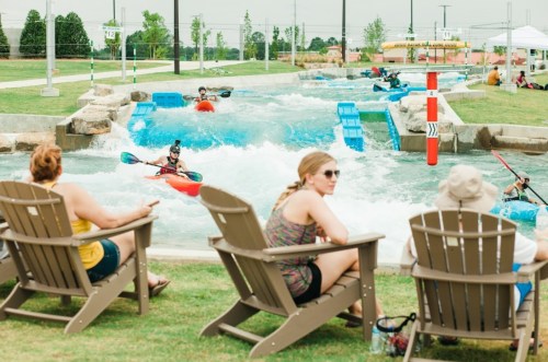People relax in chairs by a water park's wave pool, where kayakers navigate the rapids.