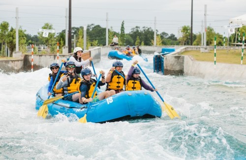 A group of people in blue rafts navigating a whitewater course, wearing life jackets and smiling.