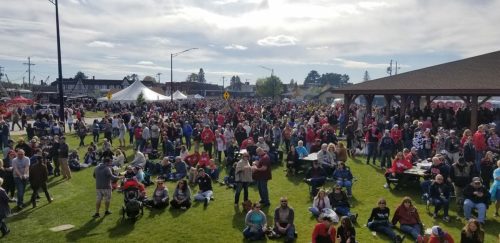 A large crowd gathers at an outdoor event, with tents and a pavilion under a partly cloudy sky.