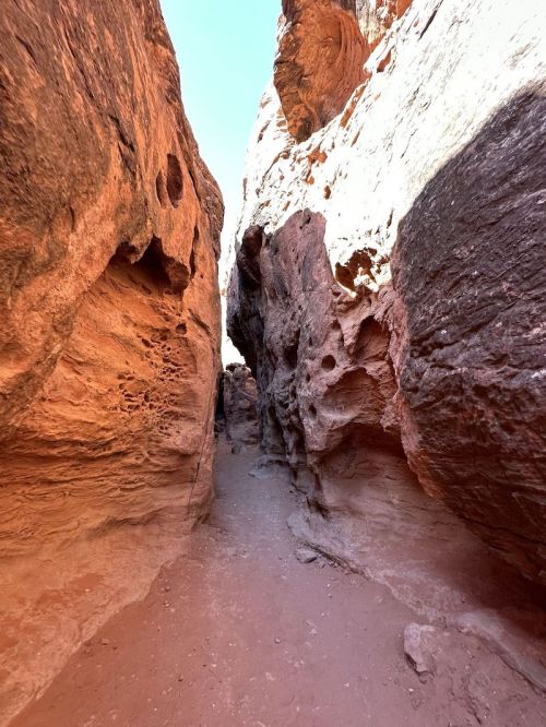 Narrow canyon with reddish rock formations and a sandy path, illuminated by bright sunlight above.