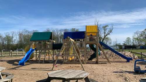 A wooden playground with slides and climbing structures, set in a sandy area under a clear blue sky.