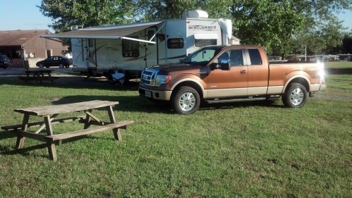 A brown and silver pickup truck parked next to a camper, with a picnic table on green grass under a clear sky.