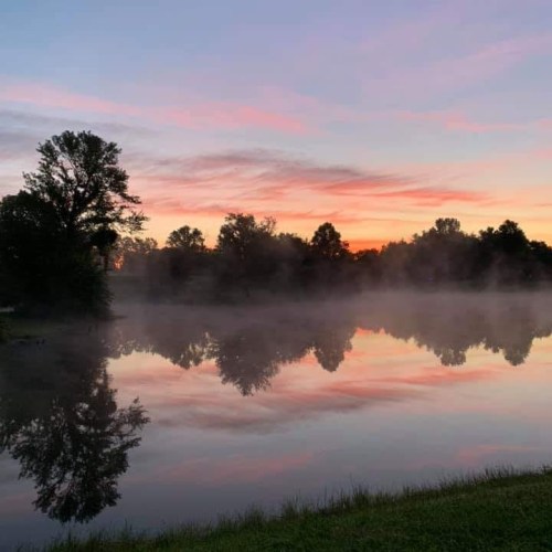 A serene lake at sunrise, with mist rising and colorful reflections of trees and clouds on the water's surface.