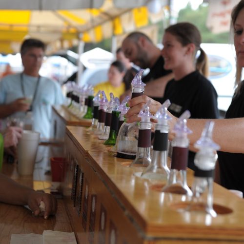 A busy outdoor bar with staff serving drinks from colorful dispensers to customers at a festival.