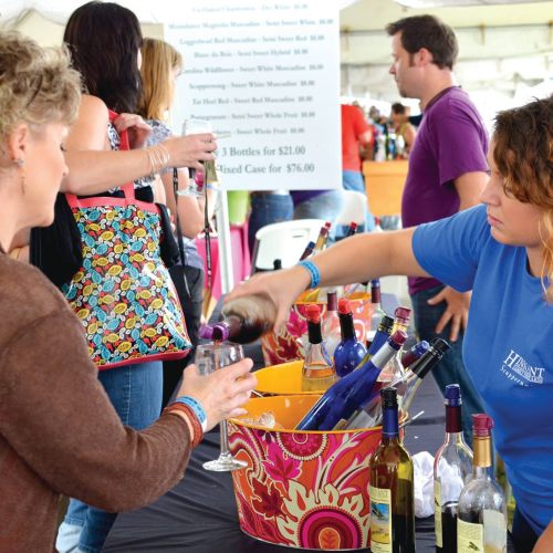 A woman pours wine into a glass for a customer at a wine tasting event, with bottles displayed on the table.