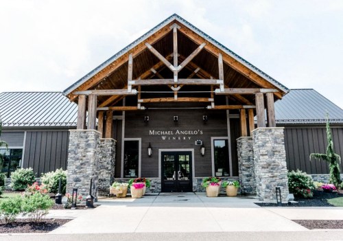 Exterior view of Michael Angelo's Winery featuring a wooden entrance and stone pillars, surrounded by colorful flower pots.