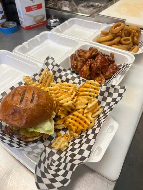A burger with lettuce, waffle fries, chicken wings, and onion rings in takeout containers on a counter.