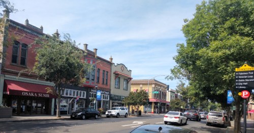 A street view featuring shops, trees, and parked cars under a clear blue sky. Signs for downtown amenities are visible.