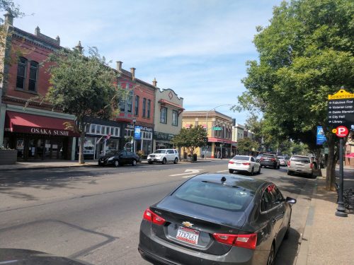 A street view featuring shops, parked cars, and trees under a clear blue sky.
