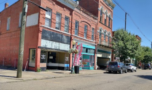 A sunny street view of brick buildings with storefronts, parked cars, and trees lining the road.