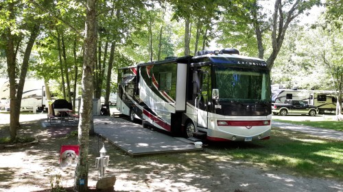 A large RV parked on a gravel site surrounded by trees, with another RV visible in the background.