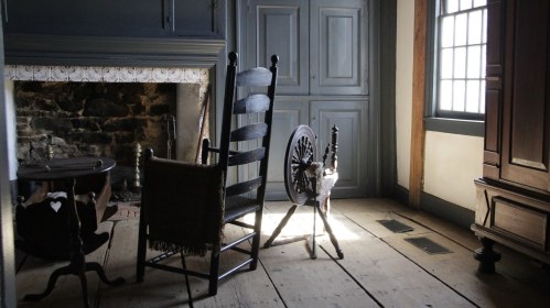 A cozy, rustic room featuring a fireplace, wooden floor, rocking chair, and a spinning wheel by a window.