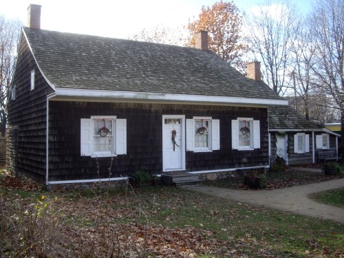 A charming, rustic house with a shingled exterior, white windows, and holiday wreaths, surrounded by autumn foliage.
