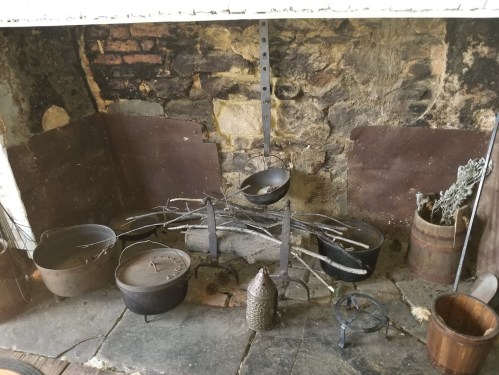 A rustic fireplace with stone walls, featuring hanging pots, a firewood rack, and various cooking utensils.