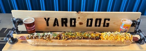 A long hot dog topped with various ingredients, served on a wooden board with two glasses of beer.