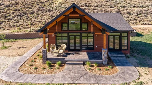 A modern cabin with large windows, a stone patio, and two chairs, surrounded by a landscaped yard and hills.