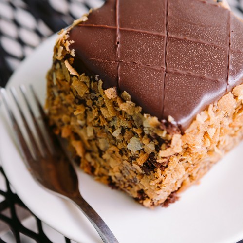 A square chocolate cake topped with a smooth chocolate layer and surrounded by crunchy flakes, served on a white plate.