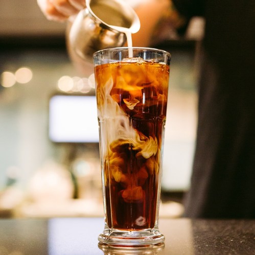 A glass of iced coffee with swirling cream being poured in, creating a marbled effect.