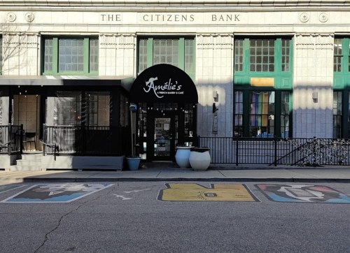 A café entrance with a black awning, colorful street art in front, and a historic bank building in the background.