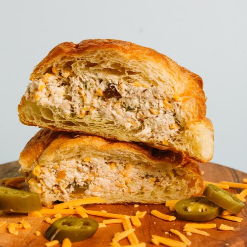 A flaky croissant sandwich filled with creamy chicken, cheese, and jalapeños, served on a wooden board.