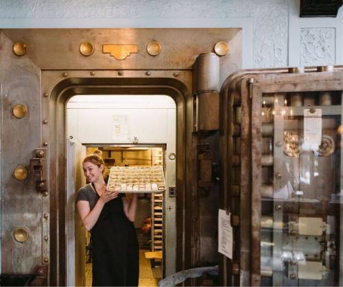 A woman holds a tray of baked goods while standing in a large, vintage-style oven door.
