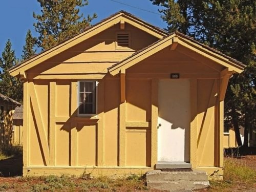 A small yellow cabin with a white door and window, surrounded by trees and blue sky.