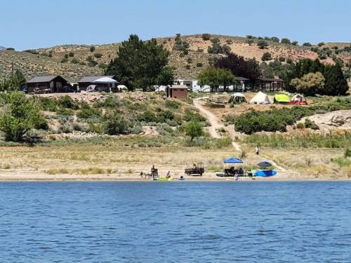 A serene lakeside scene with tents and cabins on a hillside, surrounded by greenery and a sandy beach.