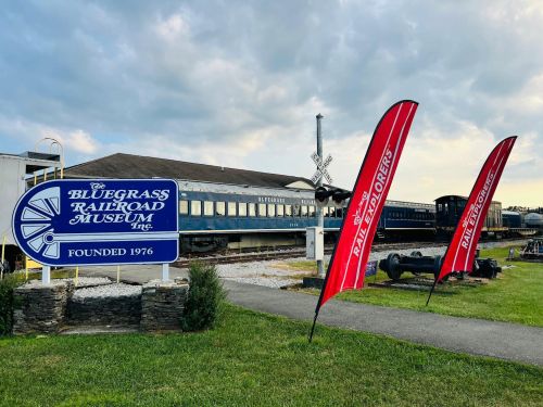 Sign for the Bluegrass Railroad Museum with flags and vintage train cars in the background under a cloudy sky.