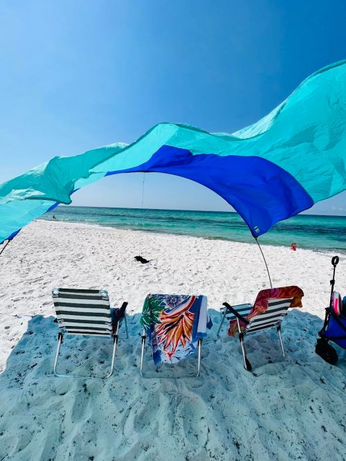 Three beach chairs under a colorful canopy, facing a sandy beach and calm ocean under a clear blue sky.