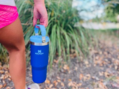 A person holds a blue Stanley tumbler while walking outdoors, surrounded by greenery and fallen leaves.