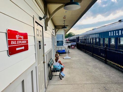 A child sits on a bench at a train station, with a sign for "Rail Explorers" and train cars in the background.