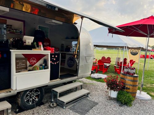 A food truck with an open side, serving drinks, surrounded by red chairs and a patio umbrella on a grassy field.