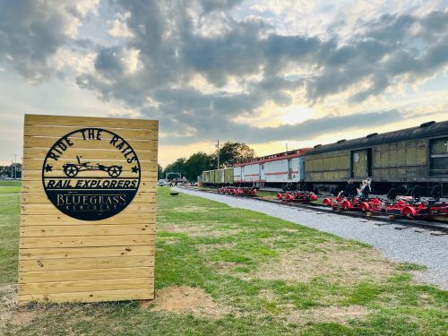 Sign for "Bluegrass Rail Explorers" with vintage train cars and pedal carts on a grassy area under a cloudy sky.