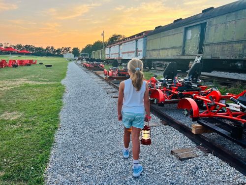 A girl walks along a gravel path holding a lantern, with go-karts and vintage train cars in the background at sunset.