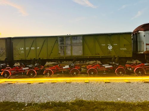 A vintage green train car beside a row of red train wheels on a gravel track at sunset.