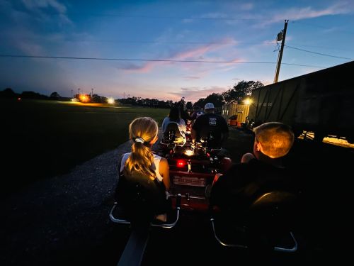 A group of people riding on a trailer at dusk, with a colorful sky and fields in the background.