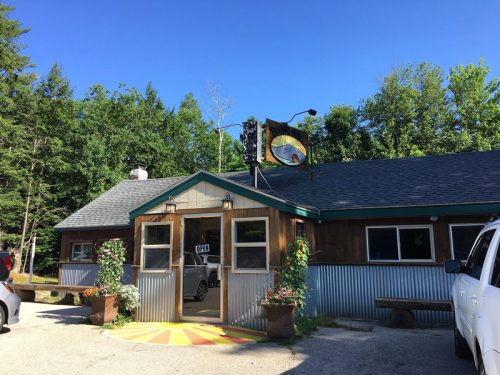 A rustic building with a sign, surrounded by trees and parked cars, under a clear blue sky.