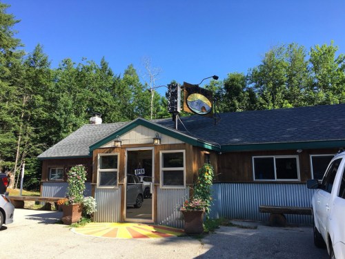 A rustic building with a sign, surrounded by trees and parked cars, under a clear blue sky.
