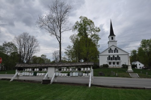 A white church with a tall steeple beside a road, featuring a sign and gravestones in the foreground under a cloudy sky.