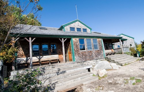 A rustic wooden cabin with a sloped roof, surrounded by greenery and blue sky. Steps lead up to the entrance.