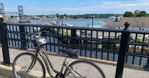 A bicycle parked by a waterfront with boats and a bridge in the background under a clear blue sky.