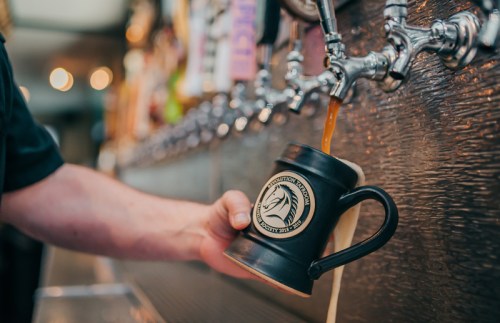 A hand pours a dark beverage from a tap into a black mug at a bar.