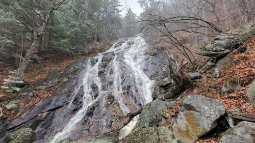 A cascading waterfall flows over rocky terrain, surrounded by trees and autumn leaves.
