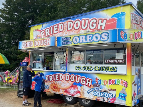 A colorful food truck selling fried dough, deep-fried Oreos, lemonade, and slushies, with two customers in front.