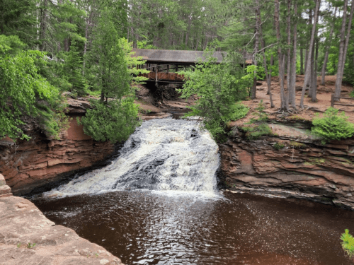 A serene waterfall cascades into a dark pool, surrounded by lush greenery and a rustic cabin in the background.