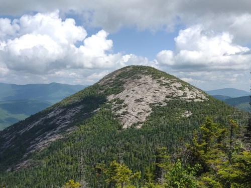 A rocky mountain peak surrounded by lush green trees and a cloudy blue sky in the background.