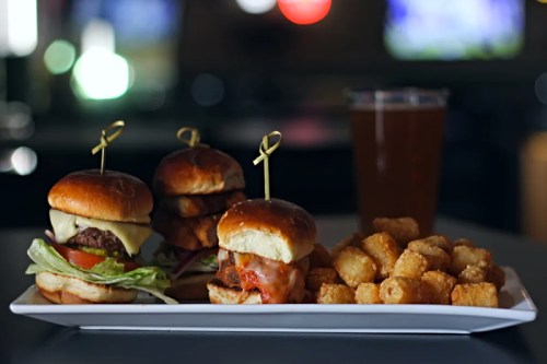 A platter with three sliders and a side of tater tots, accompanied by a glass of beer in a dimly lit setting.