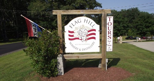 Sign for Flag Hill Winery & Distillery with a flag, listing tour times on a sunny day. Green grass and trees in background.